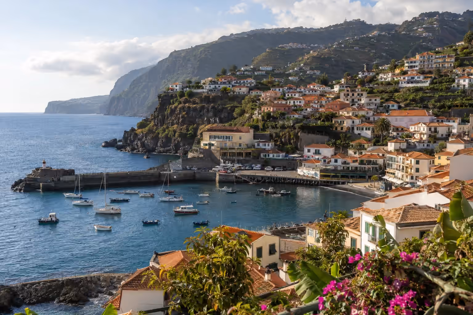 White houses with terracotta roofs on green hills above a sheltered harbour with small sailing boats