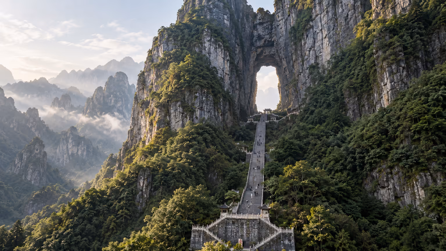 A natural rock arch above forested mountain cliffs and stone steps at Tianmen Mountain