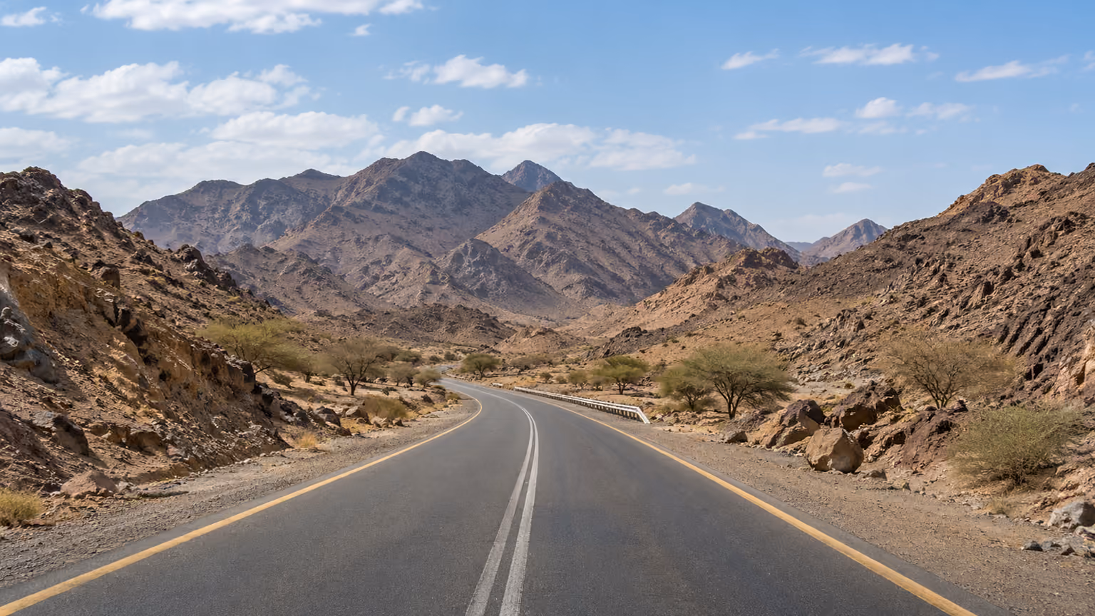 A paved road crossing a dry valley between dark rocky hills outside Madinah